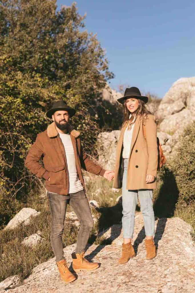 Handsome couple hiking outdoors wearing stylish casual autumn outfits, jackets and hats, holding hands on rocky trail surrounded by trees and nature, perfect for adventure travel and healthy lifestyle themes.